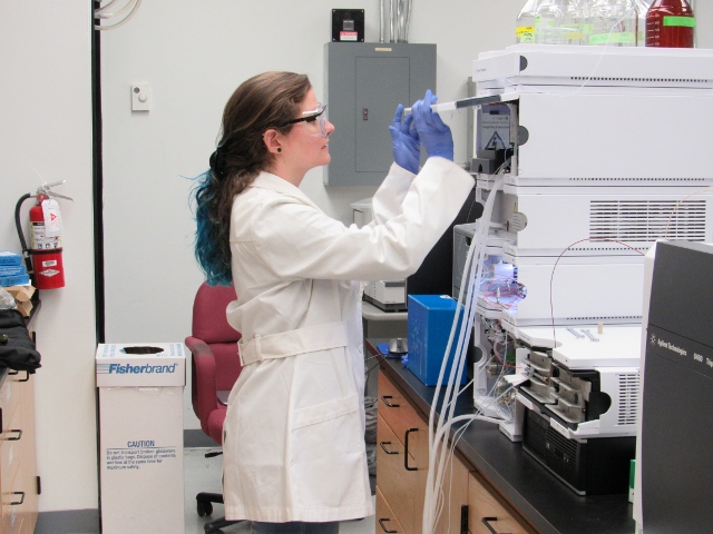 A student wears gloves, goggles, and a lab coat while working with forensic analysis equipment in a laboratory.
