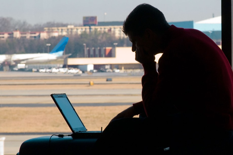 A man works at a laptop computer in an airport in front of a window overlooking the airfield and plane.